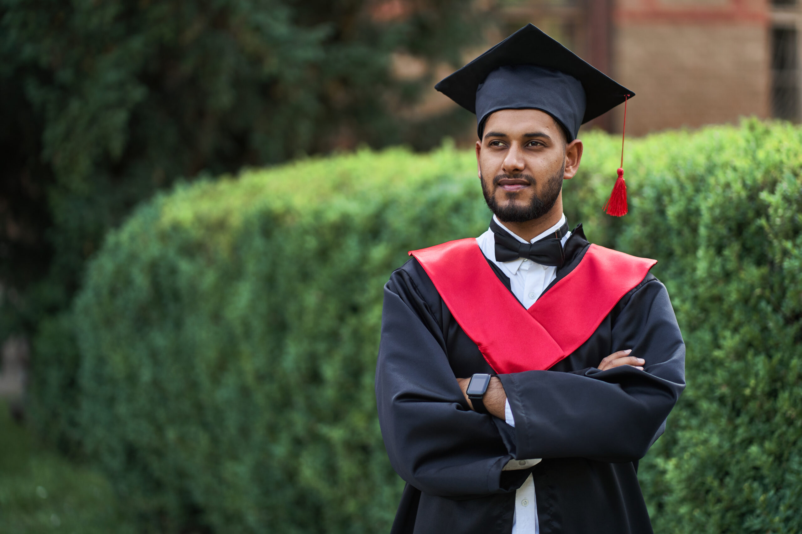 Serious indian graduate in graduation robe with crossed arms loo